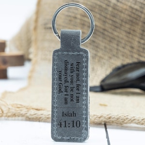 Könnte beinhalten: Ein grauer Leder-Schlüsselanhänger mit einem silbernen Schlüsselring. Auf dem Schlüsselanhänger steht der Text "Fear not, for I am with you: be not dismayed, for I am your God. Isiah 41:10". Der Hintergrund ist unscharf und neutral gefärbt.