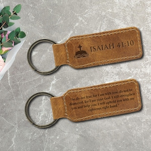 Puede incluir: Dos llaveros de cuero marrón con anillos metálicos. El llavero superior tiene el texto "ISAIAH 41:10" y una cruz sobre un libro abierto. El llavero inferior tiene un versículo bíblico. Ambos llaveros son rectangulares.