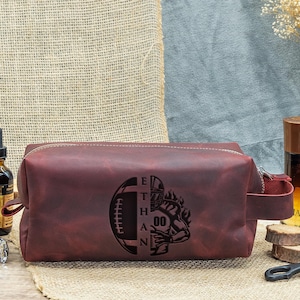 May include: A burgundy leather wash bag with a zip closure and a side handle. The bag has a football design with the name "ETHAN" and the number "00" engraved on the front.