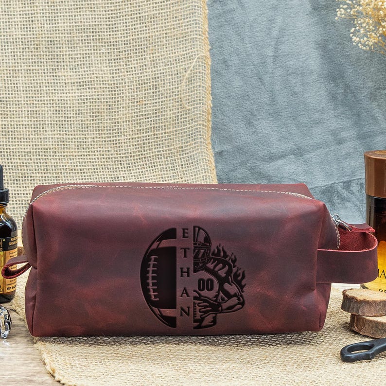 May include: A burgundy leather wash bag with a zip closure and a side handle. The bag has a football design with the name "ETHAN" and the number "00" engraved on the front.