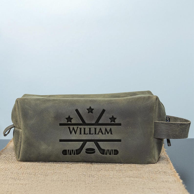 May include: A grey leather wash bag with a zip closure and a handle. The bag features a hockey-themed design with crossed hockey sticks, stars, and the name "WILLIAM" engraved on the front. The bag is on a burlap surface.