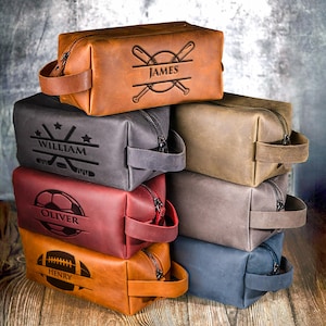 May include: A stack of personalized leather toiletry bags in various colors, including brown, gray, and red. Each bag features a zipper closure and a handle. The bags are embossed with names and sports-themed designs, such as baseball bats, hockey sticks, a soccer ball, and a football.