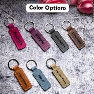 May include: Seven leather keychains with different colors and engraved text. The colors are fuchsia, purple, anthracite, dark brown, light brown, navy, and heather forest.
