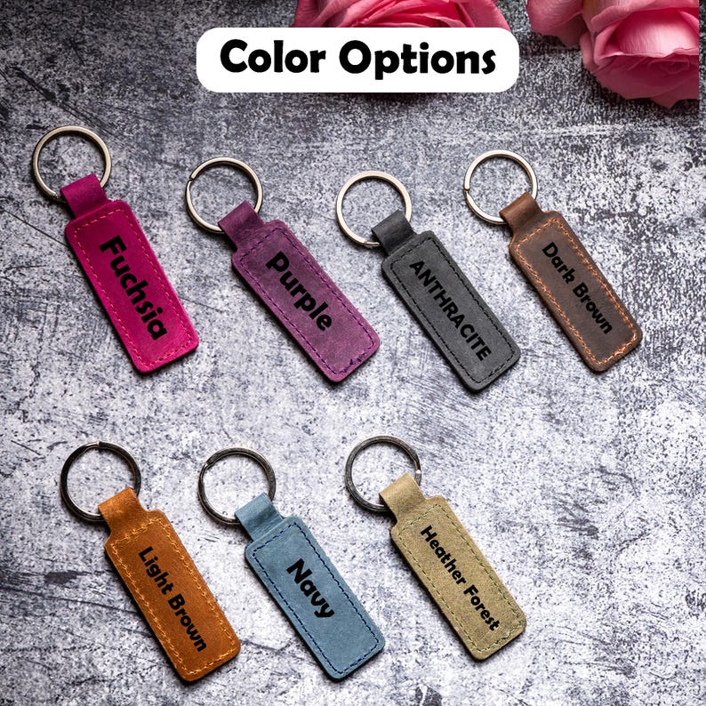 May include: Seven leather keychains with different colors and engraved text. The colors are fuchsia, purple, anthracite, dark brown, light brown, navy, and heather forest.
