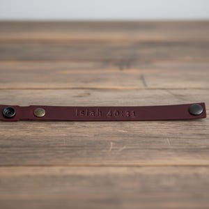 May include: A burgundy leather bracelet with the embossed text "Isaiah 40:31". The bracelet has a snap closure and a brass-colored rivet. The bracelet is displayed on a wooden surface.