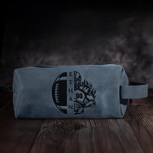 May include: A blue leather wash bag with a black zip and handle. The bag has a football and a football player graphic with the name "ETHAN" engraved on the front. The bag is rectangular and sits on a wooden surface.
