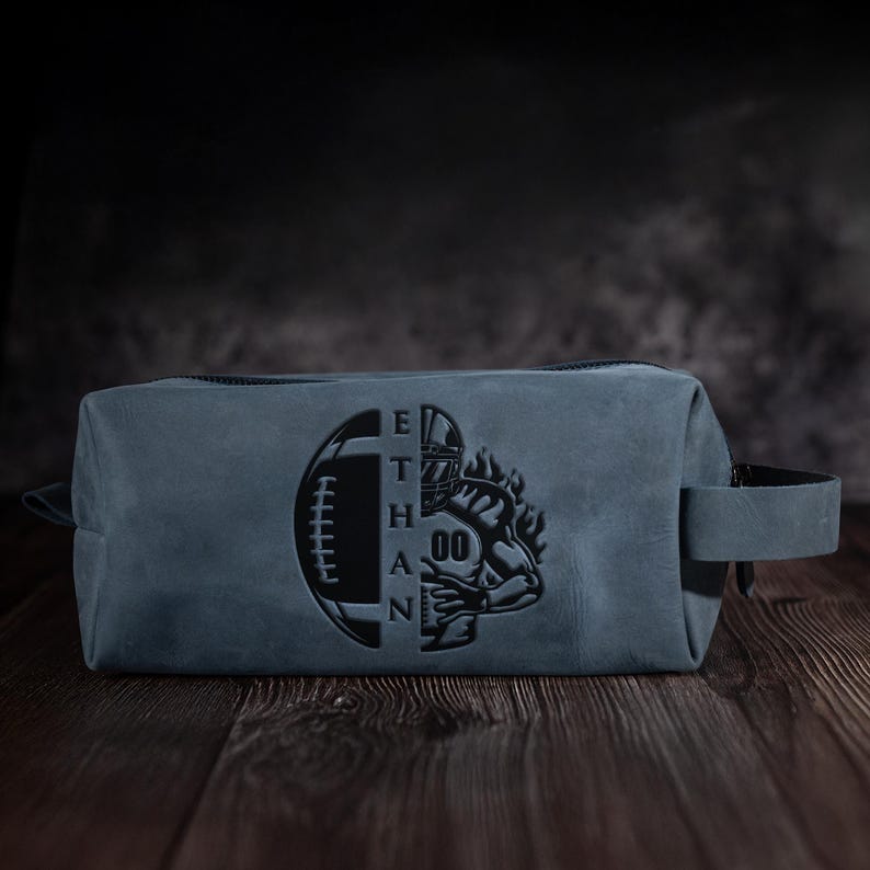 May include: A blue leather wash bag with a black zip and handle. The bag has a football and a football player graphic with the name "ETHAN" engraved on the front. The bag is rectangular and sits on a wooden surface.