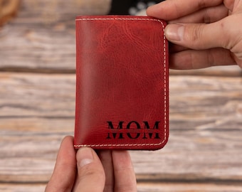 May include: A red leather wallet with the word "MOM" engraved on the front. The wallet is rectangular with rounded corners and white stitching. The wallet is held by two hands.