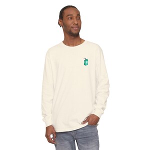 May include: Cream-colored long-sleeve shirt with a small teal juice box graphic on the chest. The shirt is made of a soft material and has a classic crew neck design. The model is wearing blue jeans.