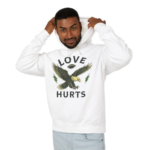 Puede incluir: Sudadera con capucha blanca con un estampado gráfico de un águila con una pelota de fútbol en sus garras. El texto "LOVE HURTS" está impreso debajo del águila.