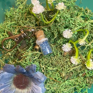 May include: A small glass bottle filled with blue glitter, attached to a silver key chain. The bottle is sitting on a bed of green moss and white flowers.