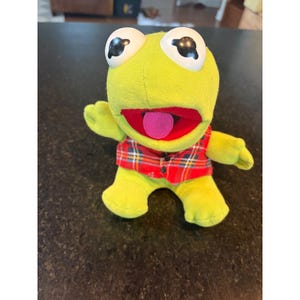 May include: A plush frog toy with large white eyes and a pink tongue. The frog is wearing a red plaid vest. The frog is lime green and has small arms and legs.