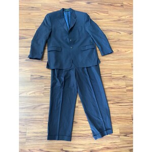 May include: A navy blue suit set, including a blazer and matching trousers. The blazer has a two-button closure and front pockets. The trousers have a pleated front and cuffed hem. The suit is made of a textured fabric and is displayed on a wooden surface.