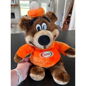 Pode incluir: Um urso de pelúcia marrom usando um chapéu e camisa laranja com o logotipo A&W. O urso tem um nariz preto, olhos brancos e uma bola branca no topo do chapéu. É um bicho de pelúcia.