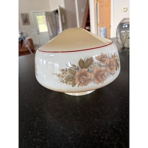 May include: A vintage-style lampshade with a cream-colored top and a white base. The base features a floral design in shades of brown and green, with a thin red stripe near the top. The lampshade is designed to sit on a base.