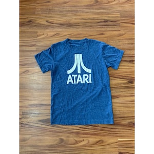 May include: Blue t-shirt with the Atari logo in white. The logo features a stylized design above the word "ATARI". The shirt is made of a soft material and has short sleeves.