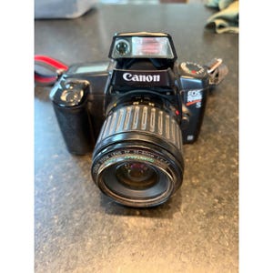 May include: Black Canon EOS Rebel S camera with a zoom lens. The camera has a built-in flash and a red strap. The lens is marked "Canon Zoom Lens EF 35-80mm 1:4-5.6".