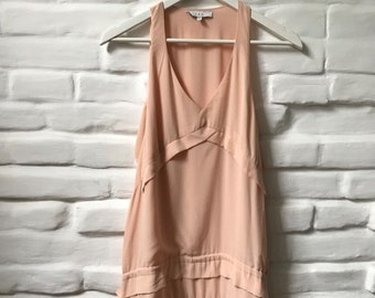 May include: A peach-colored sleeveless dress with a V-neckline and a tiered ruffle skirt. The dress is hanging on a white hanger against a white brick wall.