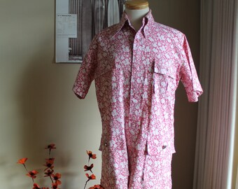 May include: A pink and white floral print short-sleeved shirt with a button-up front and two pockets. The shirt is on a mannequin and is displayed on a wooden stand.