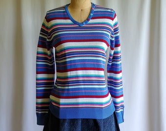 May include: A striped long-sleeved sweater with a v-neck and colorful stripes in blue, red, white, pink, and green. The sweater is on a mannequin wearing a denim skirt.