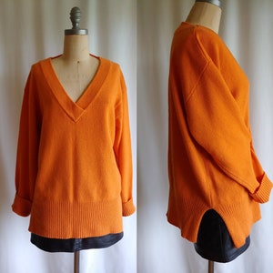 May include: A bright orange v-neck jumper with long sleeves and a ribbed hem. The jumper is made of a soft, comfortable material and has a relaxed fit.