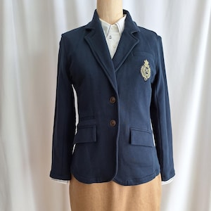 May include: A navy blue blazer with a gold embroidered crest on the left side. The blazer has a single button closure and two front pockets. The blazer is worn over a white collared shirt with a small anchor design.