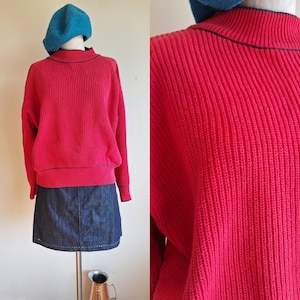 Può includere: Un maglione a coste rosso acceso con finiture nere, abbinato a una gonna di jeans e un basco blu. Il maglione ha un collo alto e maniche lunghe. La gonna è in denim scuro. L'outfit è esposto su un manichino.