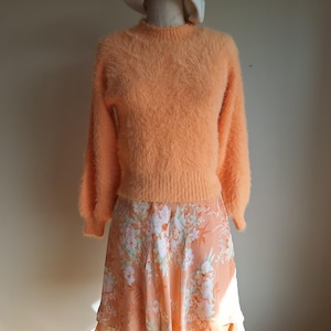 May include: A peach-coloured, fluffy jumper with a round neck and long sleeves is styled with a floral print skirt. A white beret sits on a mannequin head. The skirt has a peach background with white and green floral patterns.