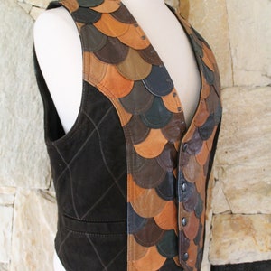 May include: A brown suede waistcoat with a unique design featuring a patchwork of leather scales in shades of brown, tan, and green. The waistcoat has a button closure and a diamond-shaped quilted pattern on the front.
