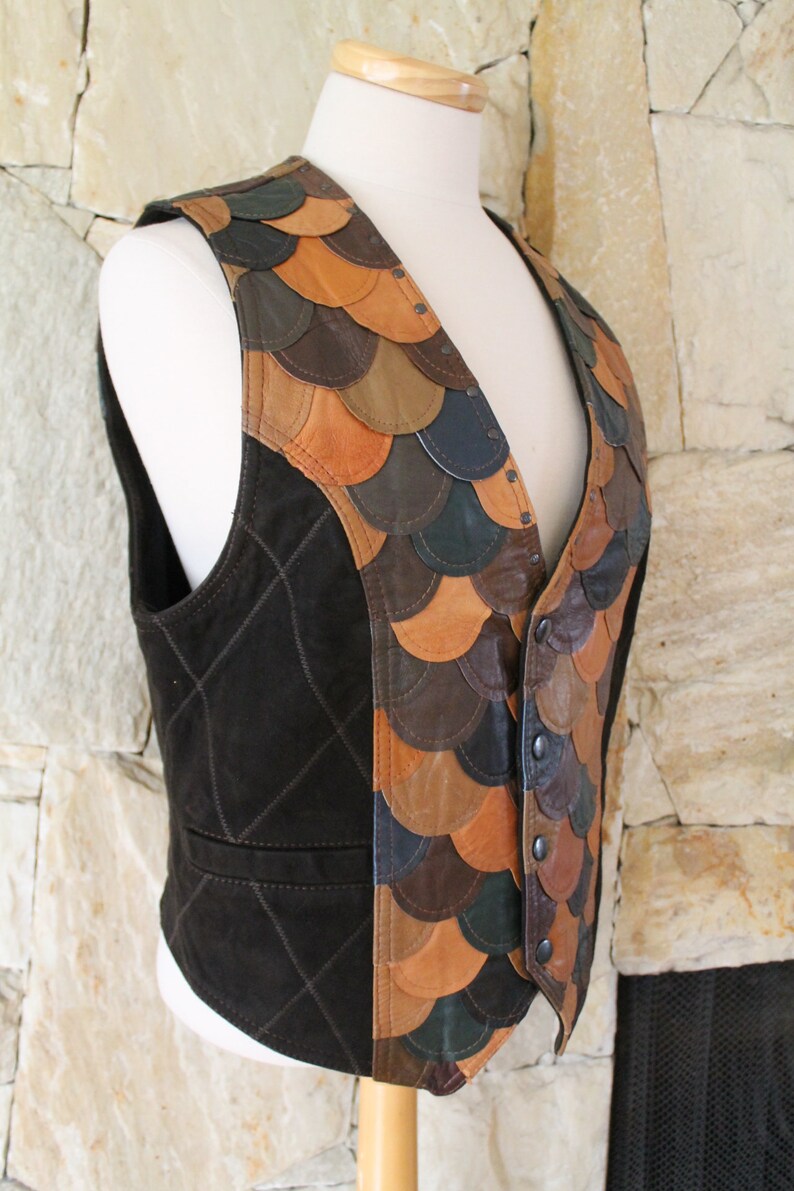 May include: A brown suede waistcoat with a unique design featuring a patchwork of leather scales in shades of brown, tan, and green. The waistcoat has a button closure and a diamond-shaped quilted pattern on the front.