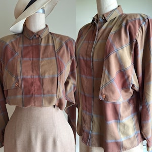 May include: A vintage-style button-down shirt in a plaid pattern of brown, tan, and muted red. The shirt has long sleeves, a collar, and a single pocket. A beige skirt is paired with the shirt. A straw hat with a black band is on the mannequin.
