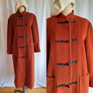 May include: A long, rust-colored wool coat with a mandarin collar and black toggle closures. The coat has two patch pockets and a relaxed fit.