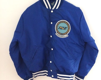 May include: A blue satin varsity jacket with white and blue striped ribbing around the collar, cuffs, and hem. The jacket has a large embroidered patch on the left chest that says "I.L.E. Electrical Workers Union District 1".