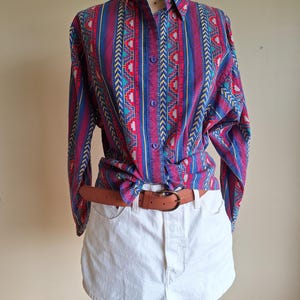 May include: A button-down shirt with a colorful geometric pattern in red, blue, and yellow is tied at the waist. It is paired with white denim shorts and a brown leather belt with a gold buckle. The shirt is displayed on a mannequin.