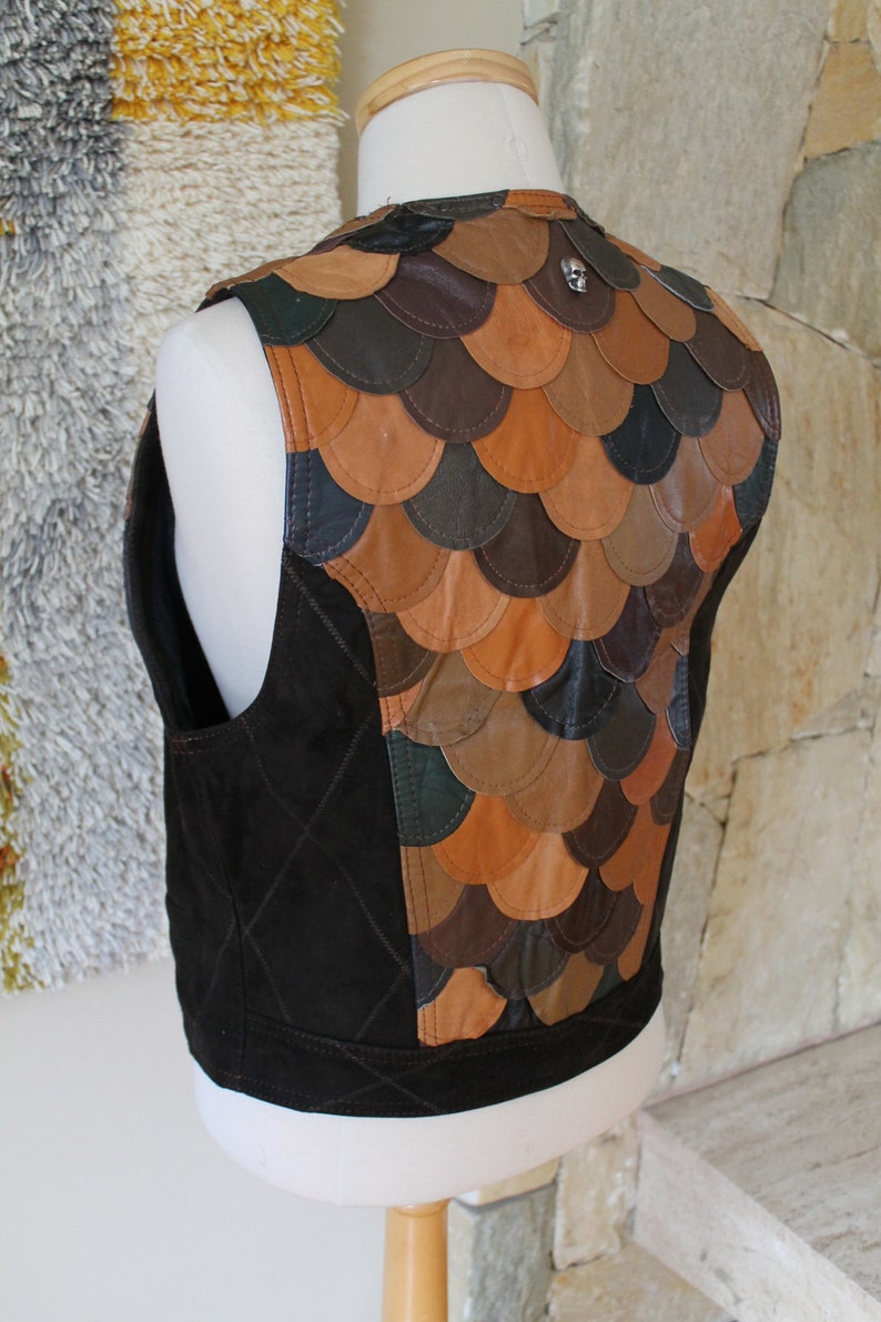 May include: A brown leather waistcoat with a quilted front and a back made of overlapping leather scales. The waistcoat has a small silver skull charm on the back.