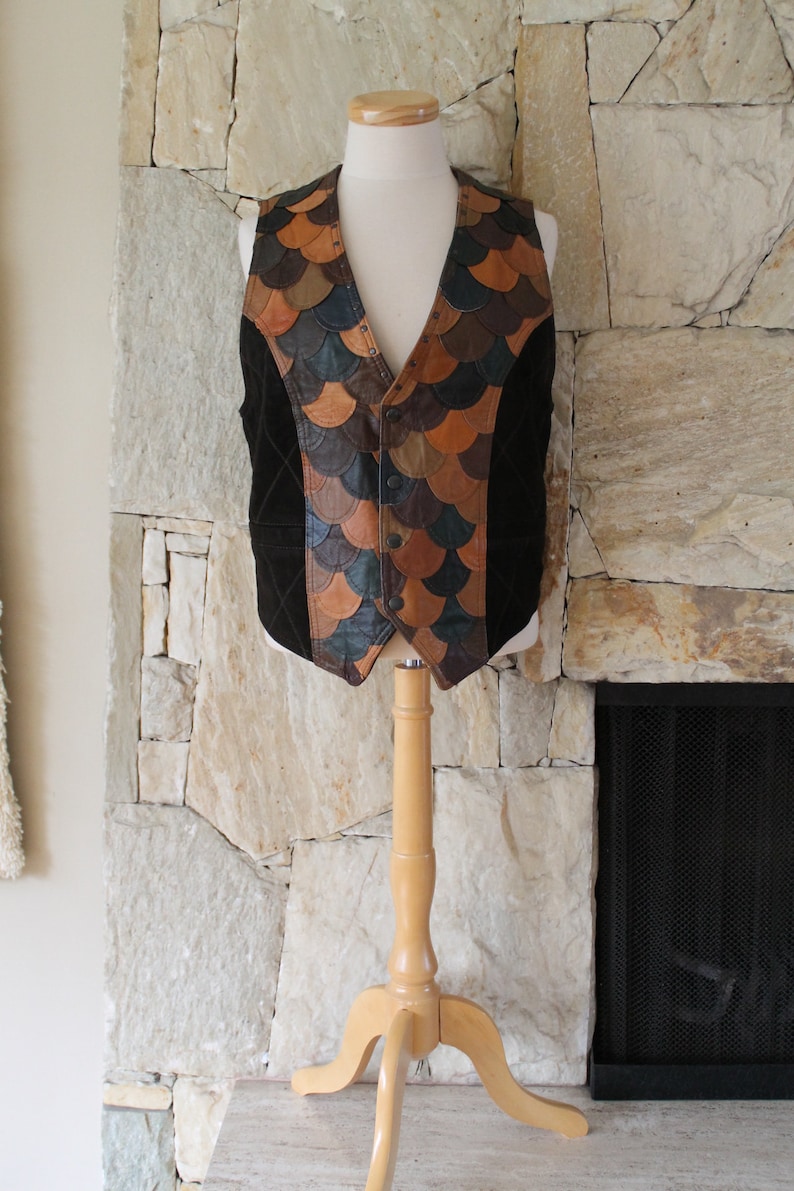 May include: A brown suede waistcoat with a patchwork design of leather pieces in shades of brown, tan, and green. The waistcoat has a button closure and is displayed on a wooden mannequin.