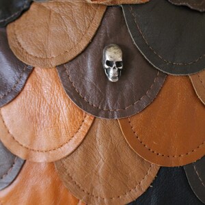 May include: A leather skirt with a scale-like design in shades of brown, tan, and black. A silver skull charm is attached to the skirt.