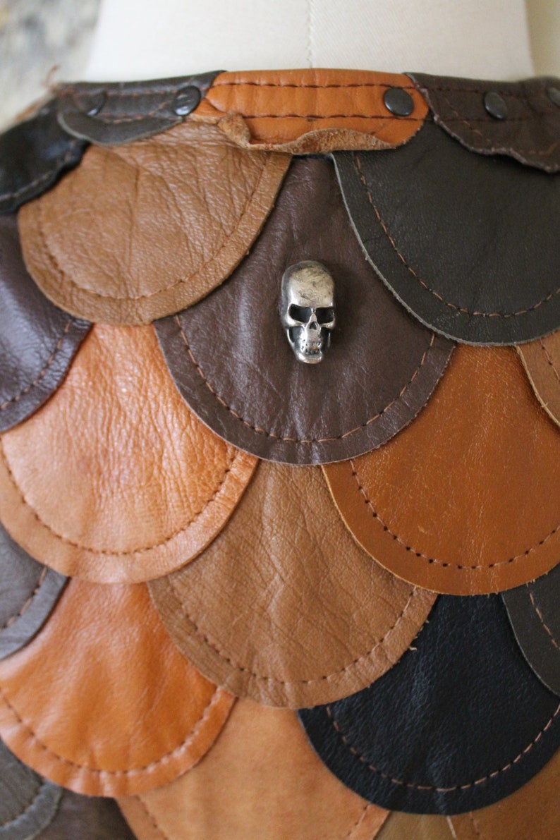May include: A leather skirt with a scale-like design in shades of brown, tan, and black. A silver skull charm is attached to the skirt.