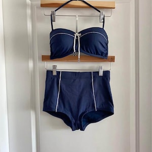 May include: Navy blue bikini set with white detailing. The bandeau-style top features a white drawstring. The high-waisted bottoms have white vertical stripes. The set is displayed on a wooden hanger.