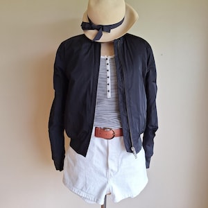 Può includere: Un ensemble di moda con un bomber nero, pantaloncini di jeans bianchi e una maglia a righe. Un cappello beige con un nastro nero e una cintura marrone completano l'outfit. L'abbigliamento è esposto su un manichino.