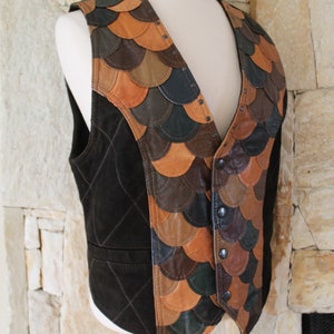 May include: A brown suede waistcoat with a quilted pattern and a decorative front panel made of various shades of brown, tan, and green leather. The leather pieces are sewn together in a scale-like pattern.