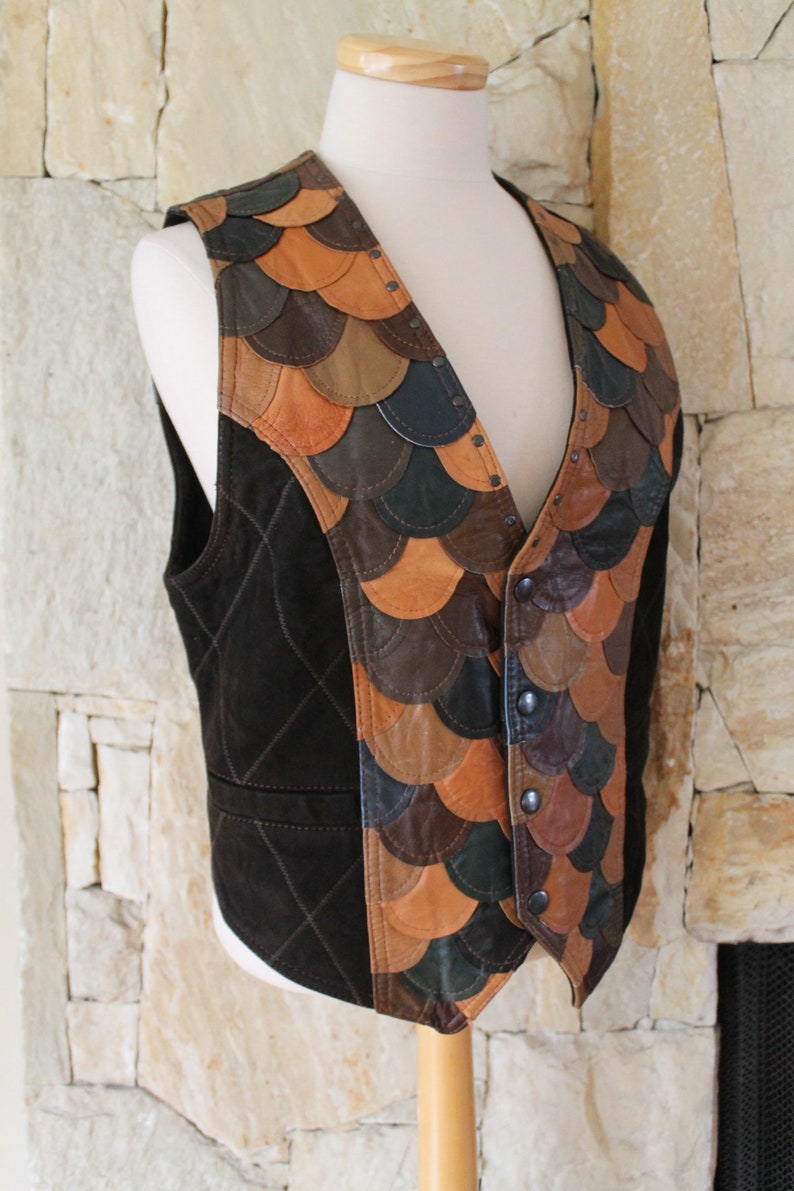 May include: A brown suede waistcoat with a quilted pattern and a decorative front panel made of various shades of brown, tan, and green leather. The leather pieces are sewn together in a scale-like pattern.
