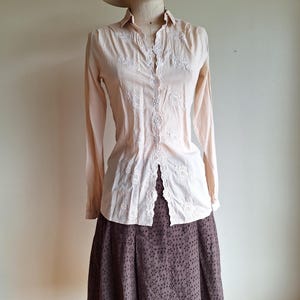 May include: A pale peach long-sleeved button-down shirt featuring white floral embroidery along the front placket and collar, paired with a dark brown eyelet skirt and a beige sun hat with a black ribbon band displayed on a mannequin.