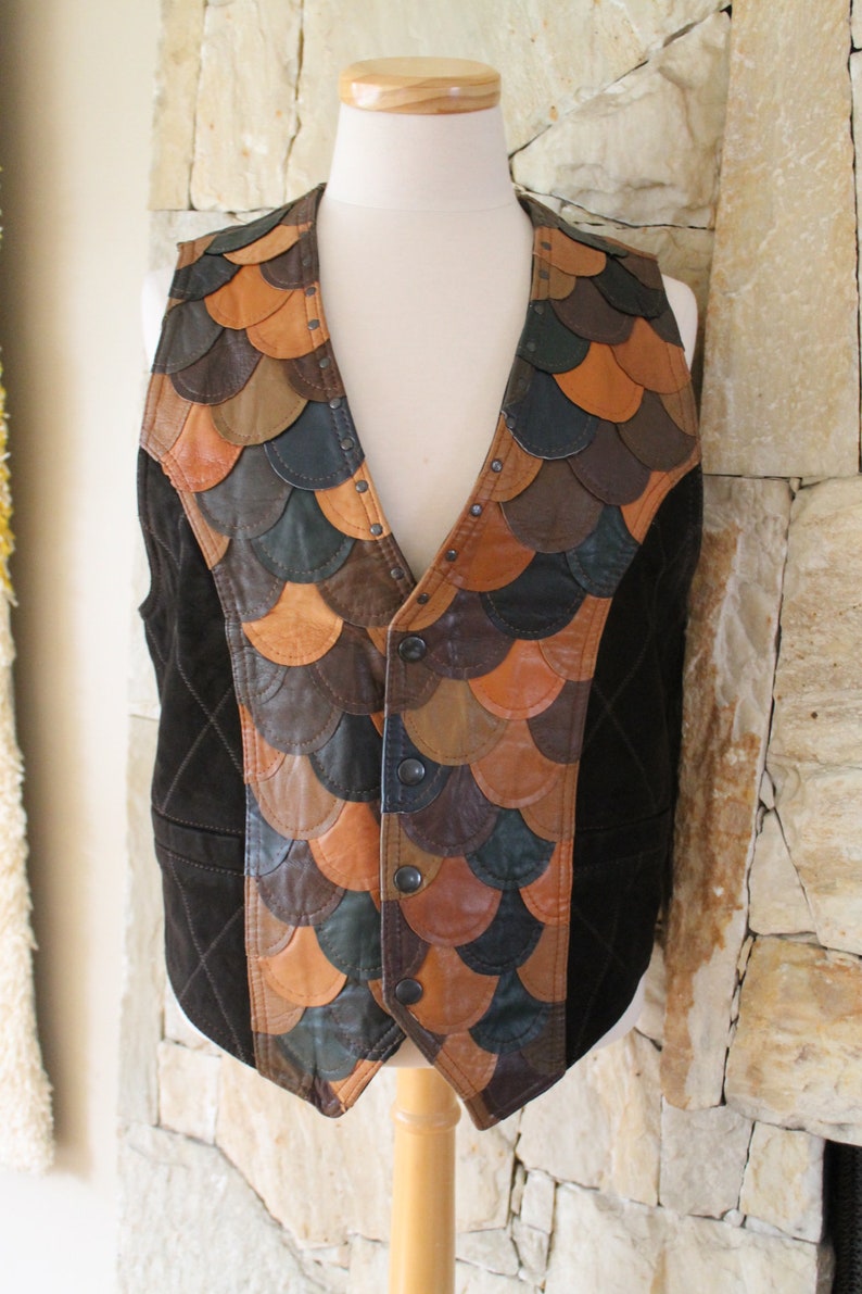 May include: A brown suede waistcoat with a unique design featuring a patchwork of leather scales in shades of brown, tan, and black. The waistcoat has a button closure and is accented with metal studs.
