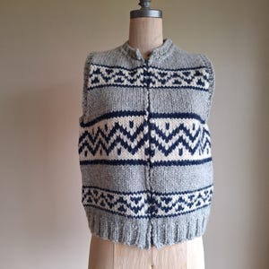 May include: A sleeveless knit waistcoat featuring a grey base with a repeating navy blue and cream geometric zigzag pattern. The garment has a front zip closure, a ribbed hem, and a high neckline, constructed from a thick, textured wool-like yarn.