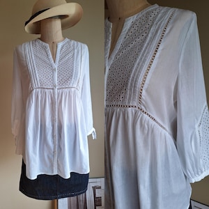 Puede incluir: Blusa blanca de manga larga con detalles calados y plisados. La blusa tiene cuello en V y cierre de botones en la parte delantera. Las mangas están recogidas en los puños. Un sombrero de paja con una banda negra está en la cabeza del maniquí.
