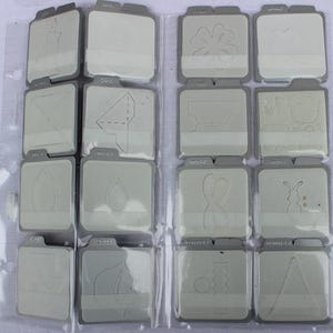 May include: A close-up of a white album with 16 clear plastic pockets, each containing a different die-cut shape. The shapes include a candle, a clover, a leaf, a heart, a caterpillar, and a triangle.