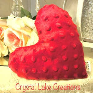May include: A red heart-shaped pillow with a textured surface. The pillow is made of soft, plush fabric and has a subtle sheen. The pillow is sitting on a white surface with a lace overlay.