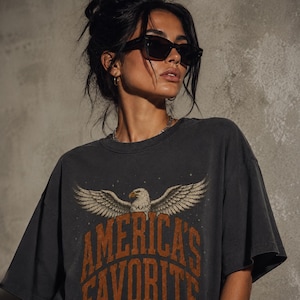 May include: Dark gray t-shirt featuring an eagle graphic with outstretched wings and the text "America's Favorite" in orange. The shirt has a vintage, worn look and a relaxed fit. The shirt is a casual wear item.