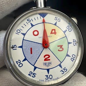 Może przedstawiać: Srebrny stoper z białą tarczą i czerwonymi, niebieskimi i zielonymi sekcjami. Tarcza ma czarne liczby od 10 do 55 oraz nadrukowane słowa "Yacht Timer" i "Heuer". Stoper ma czerwoną wskazówkę wskazującą na cyfrę 4.
